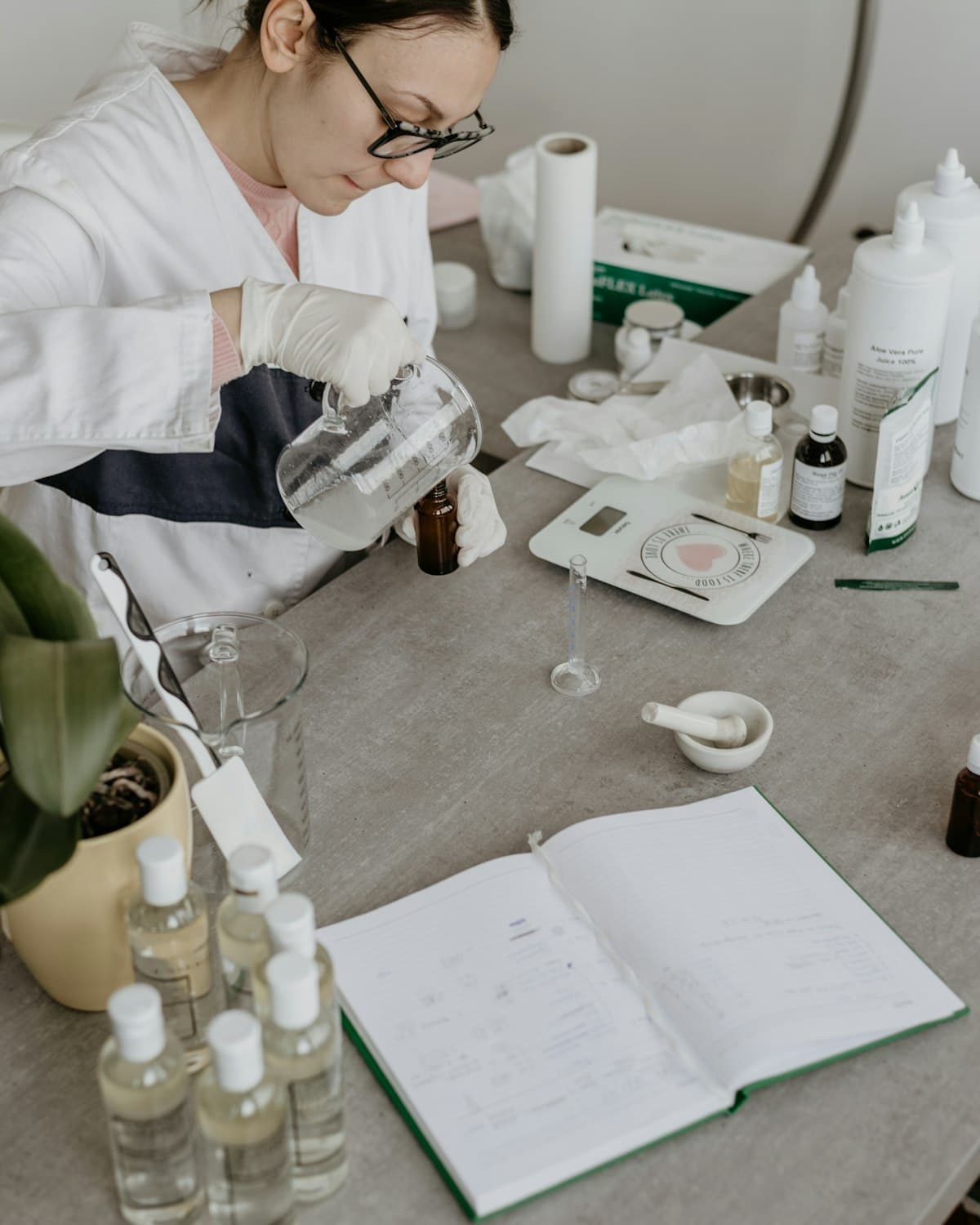 Formulator perempuan berjas laboratorium menuang bahan aktif ke dalam botol, dikelilingi bahan baku dan catatan formula